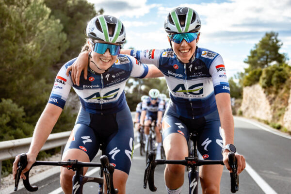 Twee wielrenners van de vrouwenploeg AG Insurance Soudal Quickstep omarmen elkaar tijdens het fietsen.